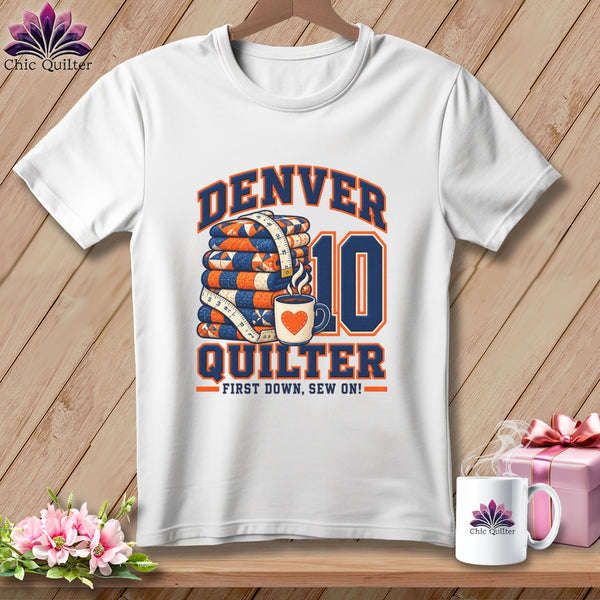 A white t-shirt hanging against a wooden wall features graphic text reading Denver Quilter with a stack of quilts, a measuring tape, a coffee cup, and the number ten.