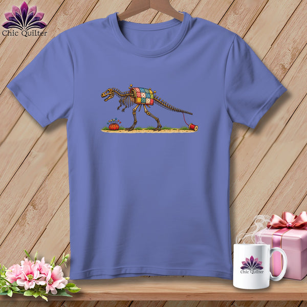 A periwinkle t-shirt featuring an illustration of a dinosaur skeleton with a patchwork quilt body on a wooden background.