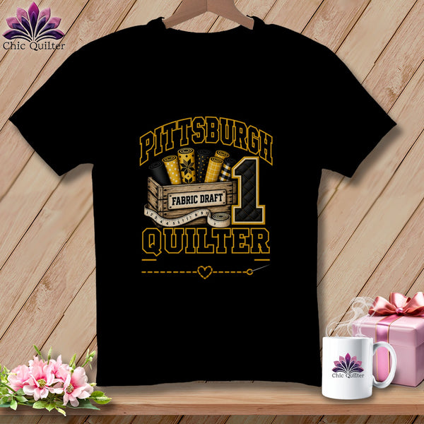 A black short-sleeve t-shirt features a graphic with yellow text reading Pittsburgh Quilter, a wooden crate of fabric rolls, a measuring tape, and a number one.