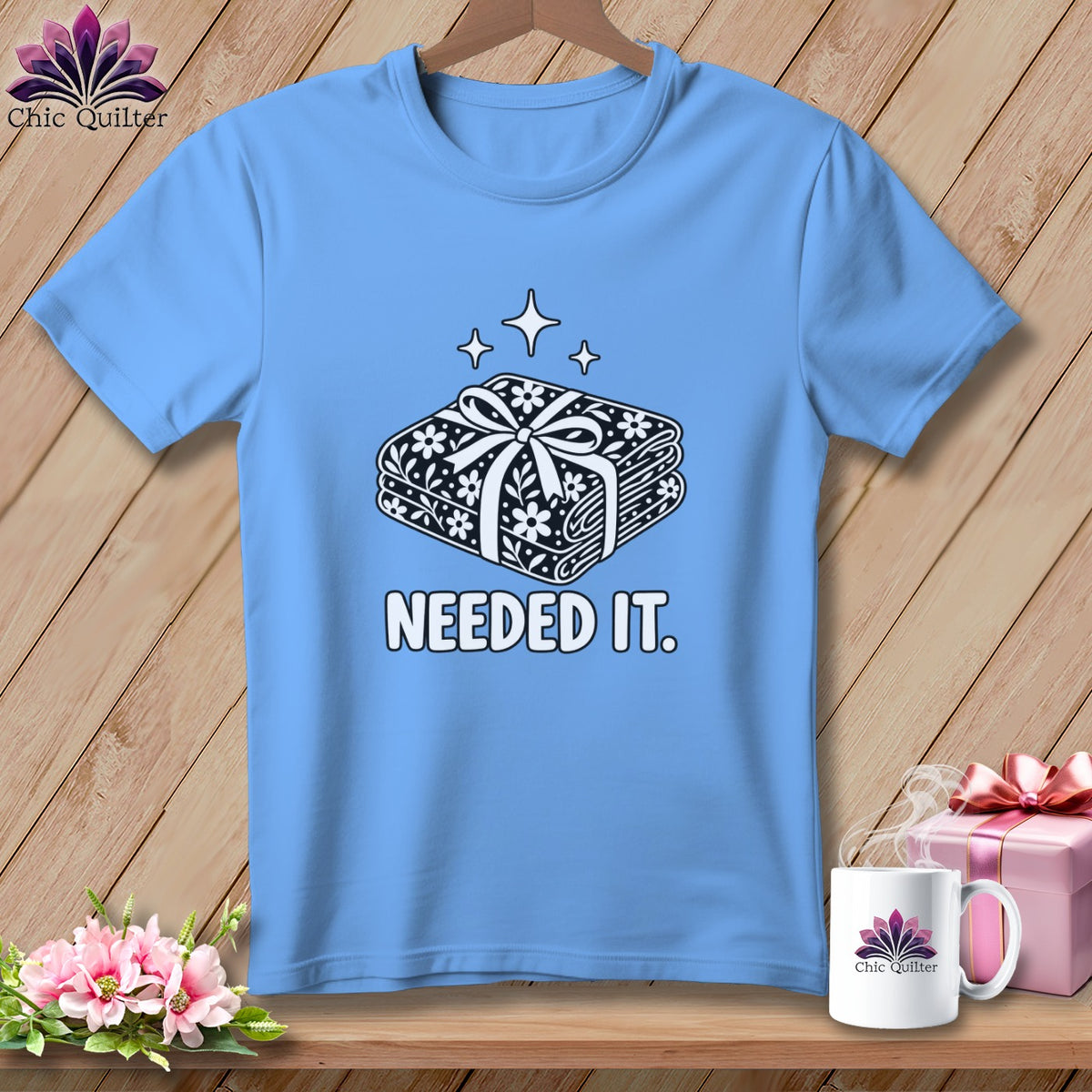 Light blue t-shirt with a stack of folded fabric tied with a bow and "NEEDED IT." text.