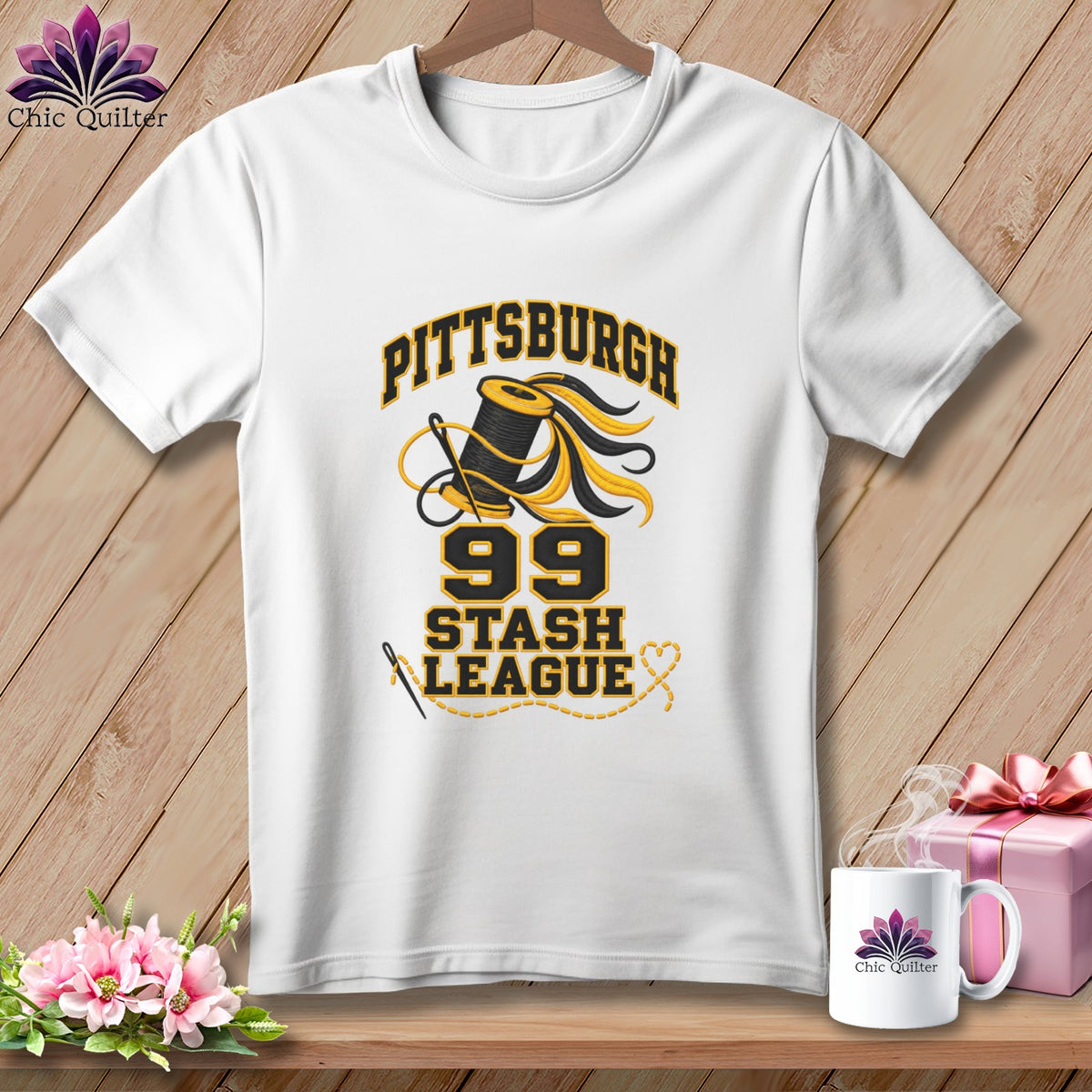 A white short-sleeved t-shirt hangs on a hanger, featuring black and yellow text reading Pittsburgh 99 Stash League alongside a spool of thread and a needle.