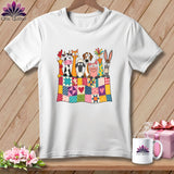 A white t-shirt hanging on a wooden background, featuring a graphic of farm animals peeking over a colorful patchwork quilt.