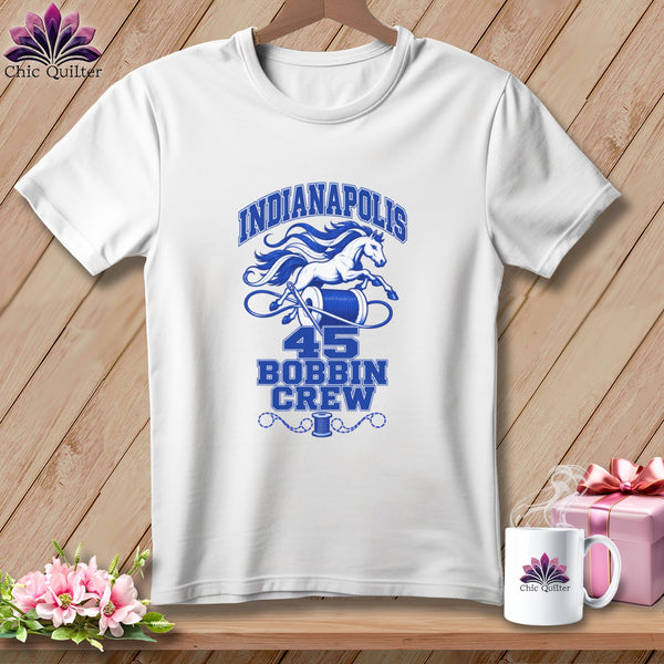 A white t-shirt hanging on a wooden wall features a blue graphic with a horse, a spool of thread, and text that reads Indianapolis 45 Bobbin Crew.