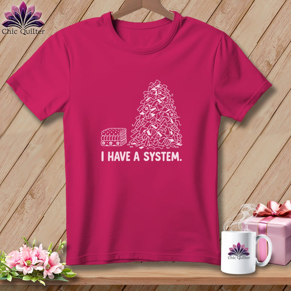 Pink t-shirt with a stack of folded fabric and chaotic fabric scraps, text "I HAVE A SYSTEM."