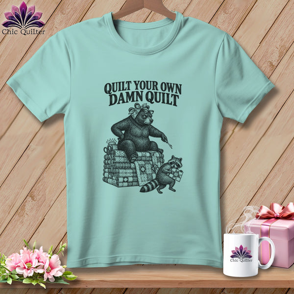 A mint green t-shirt featuring a black graphic of a bear and raccoon sewing with the text Quilt Your Own Damn Quilt.