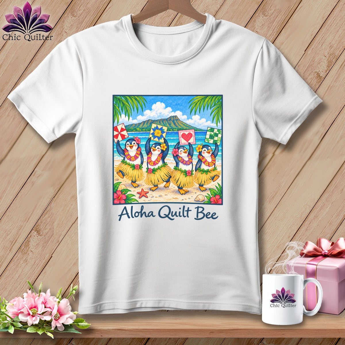 A white t-shirt hanging against a wooden wall features a graphic of four hula-dancing penguins on a beach, with the text Aloha Quilt Bee underneath.