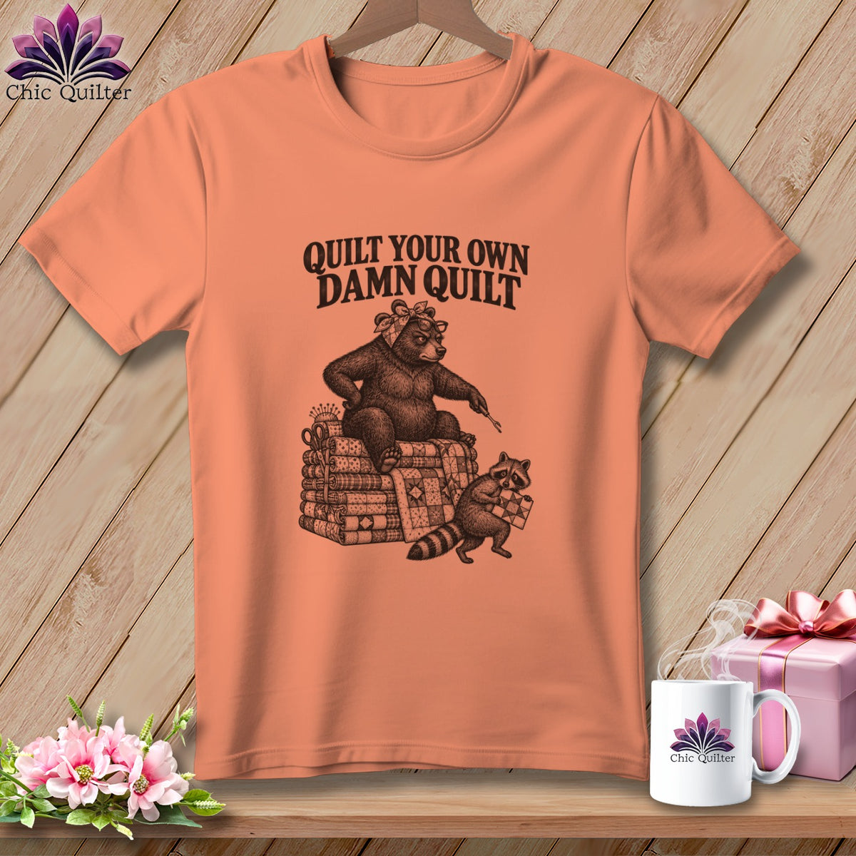 Peach short-sleeve t-shirt with a bear and raccoon graphic and the text Quilt Your Own Damn Quilt.