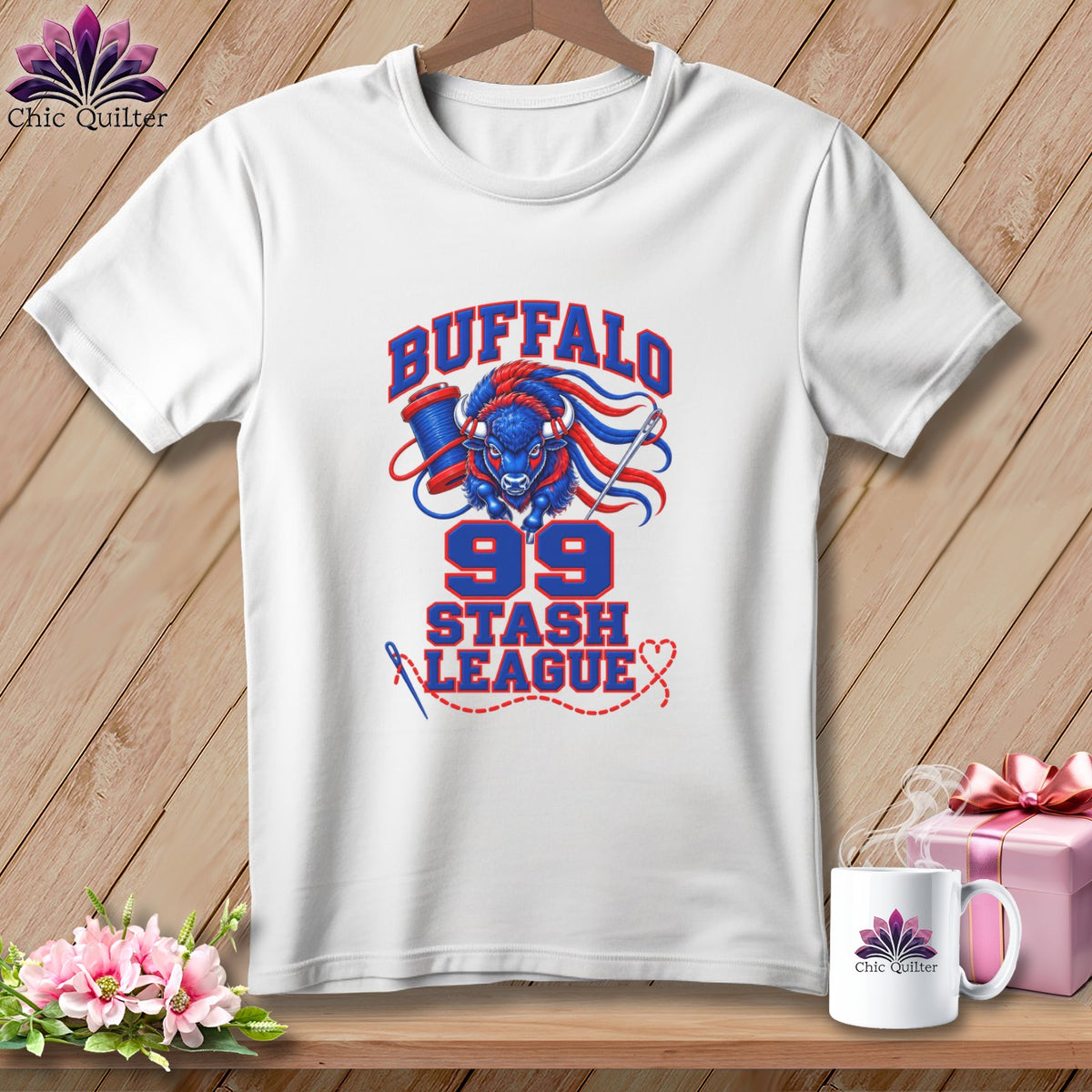 A white t-shirt hanging on a wooden background, featuring a blue and red graphic with a buffalo, sewing spool, needle, and text that reads "Buffalo 99 Stash League".