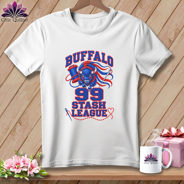 A white t-shirt hanging on a wooden background, featuring a blue and red graphic with a buffalo, sewing spool, needle, and text that reads "Buffalo 99 Stash League".