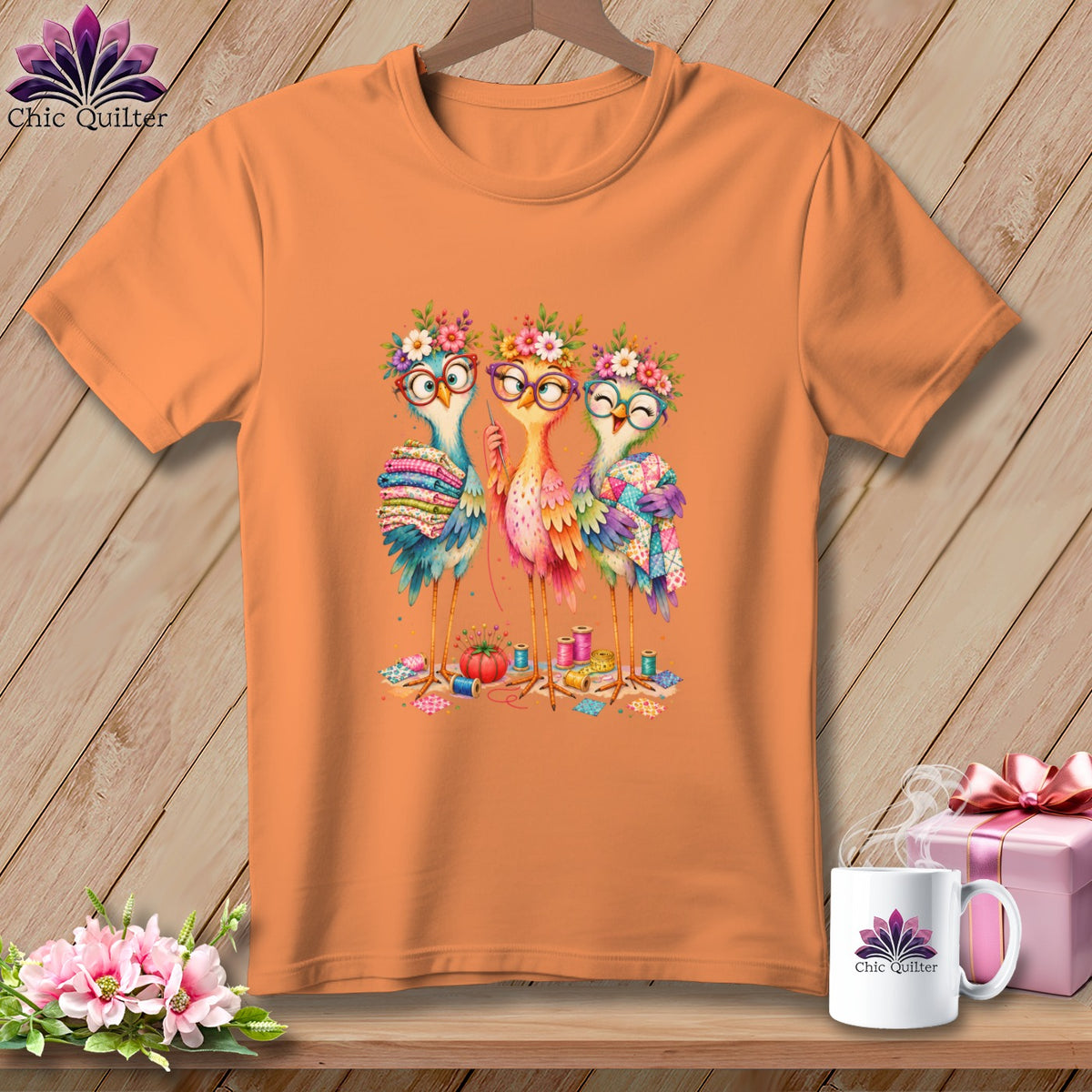An orange crewneck t-shirt featuring a cartoon illustration of three colorful birds sewing and crafting.