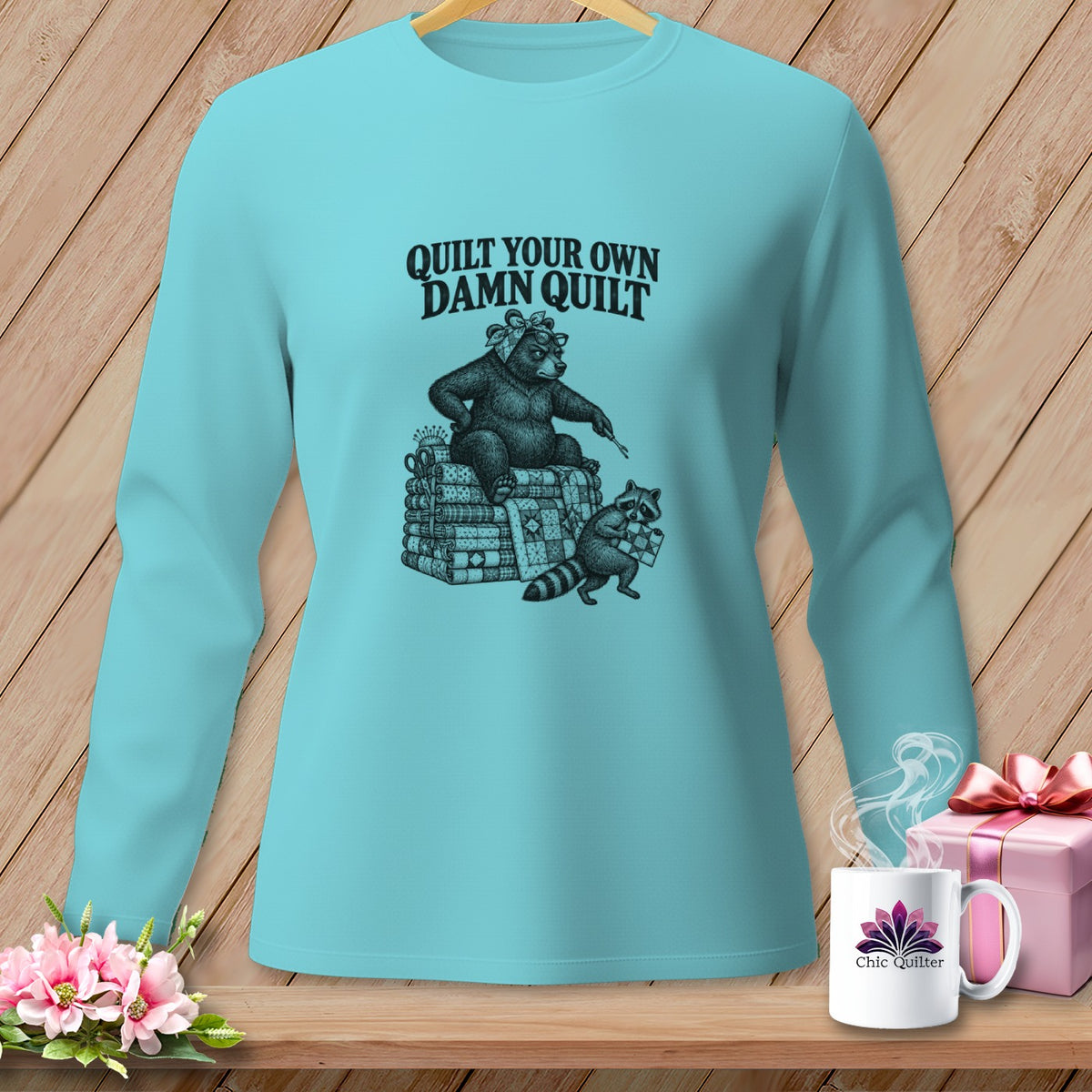 Light blue long-sleeve t-shirt with a graphic of a bear and raccoon quilting and the text Quilt Your Own Damn Quilt.