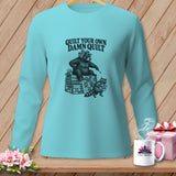 Light blue long-sleeve t-shirt with a graphic of a bear and raccoon quilting and the text Quilt Your Own Damn Quilt.