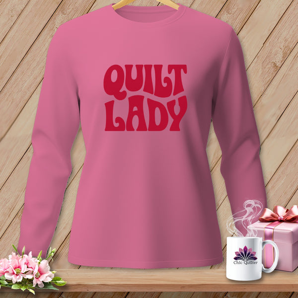 Pink long sleeve shirt with "Quilt Lady" text in red, on display with a mug and gift.