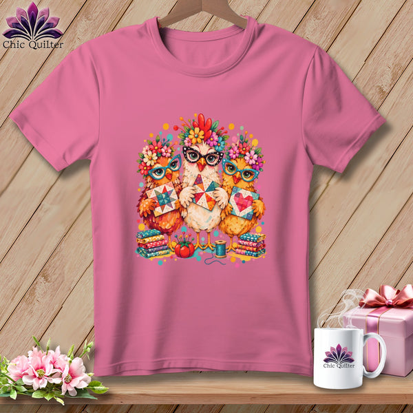 A pink crewneck t-shirt featuring a graphic of three chickens wearing flower crowns and glasses, holding quilt blocks.