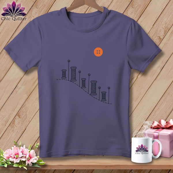 Purple t-shirt with a graphic of spools of thread, pins, and an orange button.
