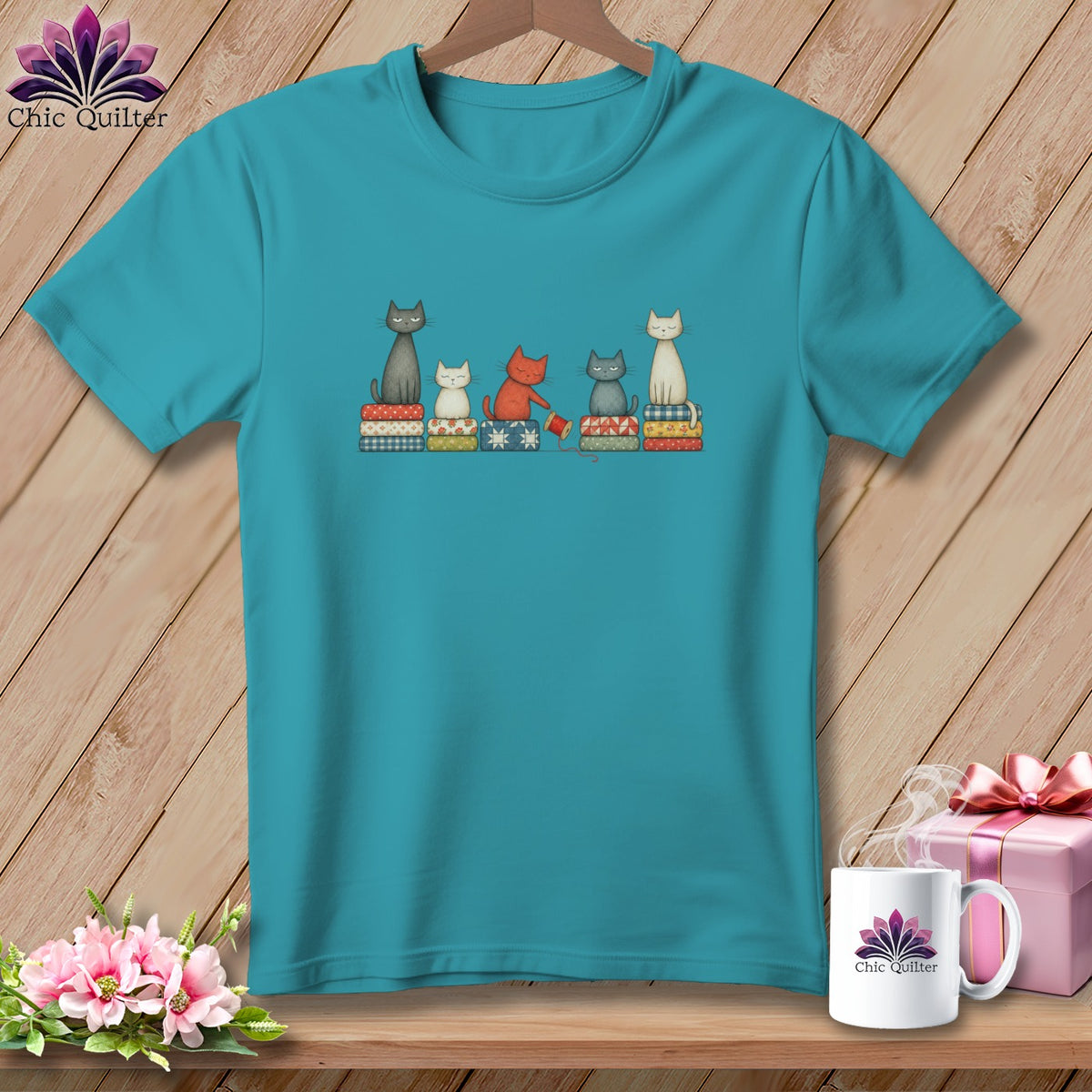 Teal crewneck t-shirt featuring a graphic of five illustrated cats sitting on stacks of colorful quilts.