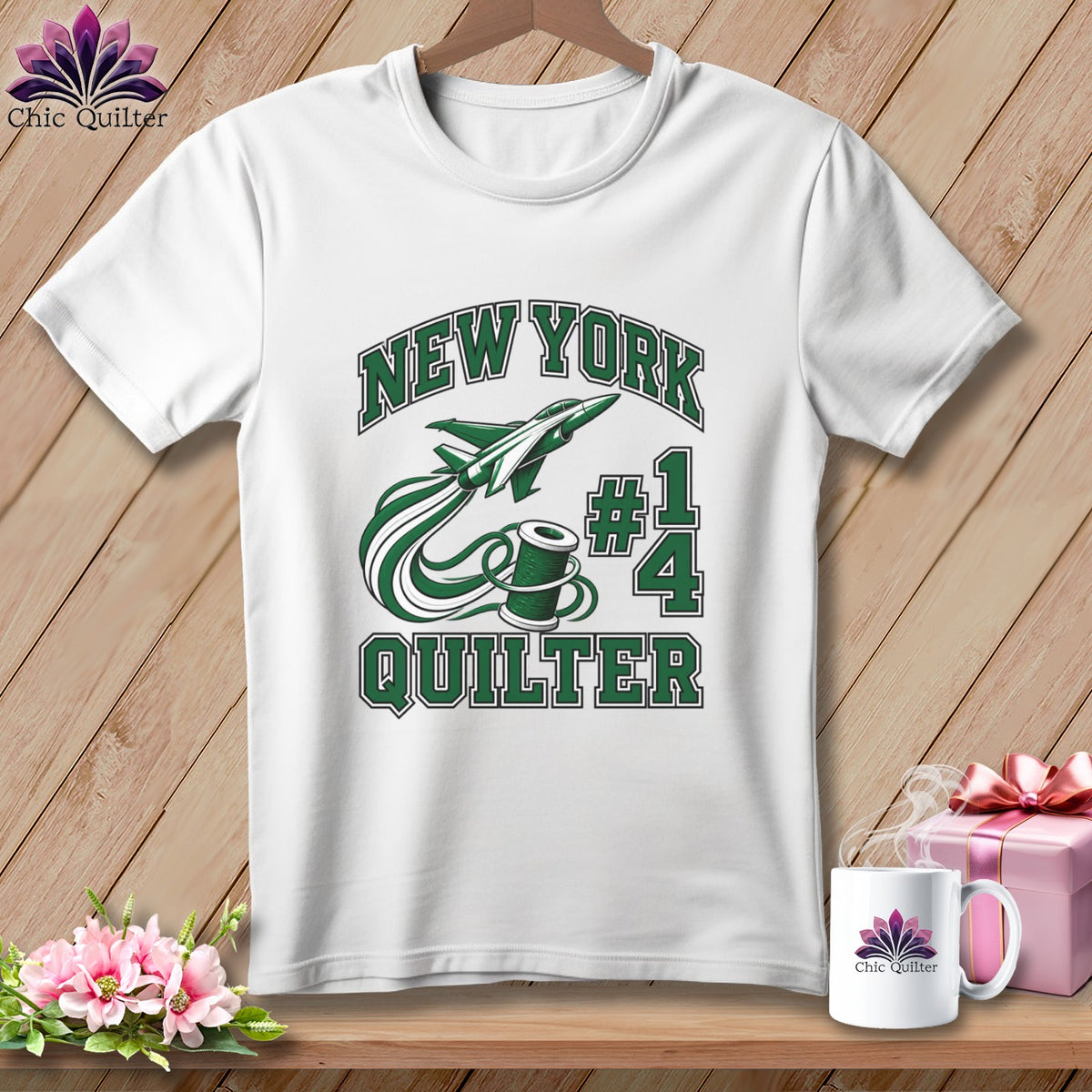 A white t-shirt hanging on a wooden background, featuring green text that reads New York Quilter, a spool of thread, and a jet plane.
