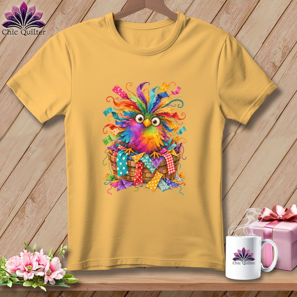 Gold t-shirt featuring a colorful, whimsical owl illustration sitting inside a woven basket of fabric scraps.