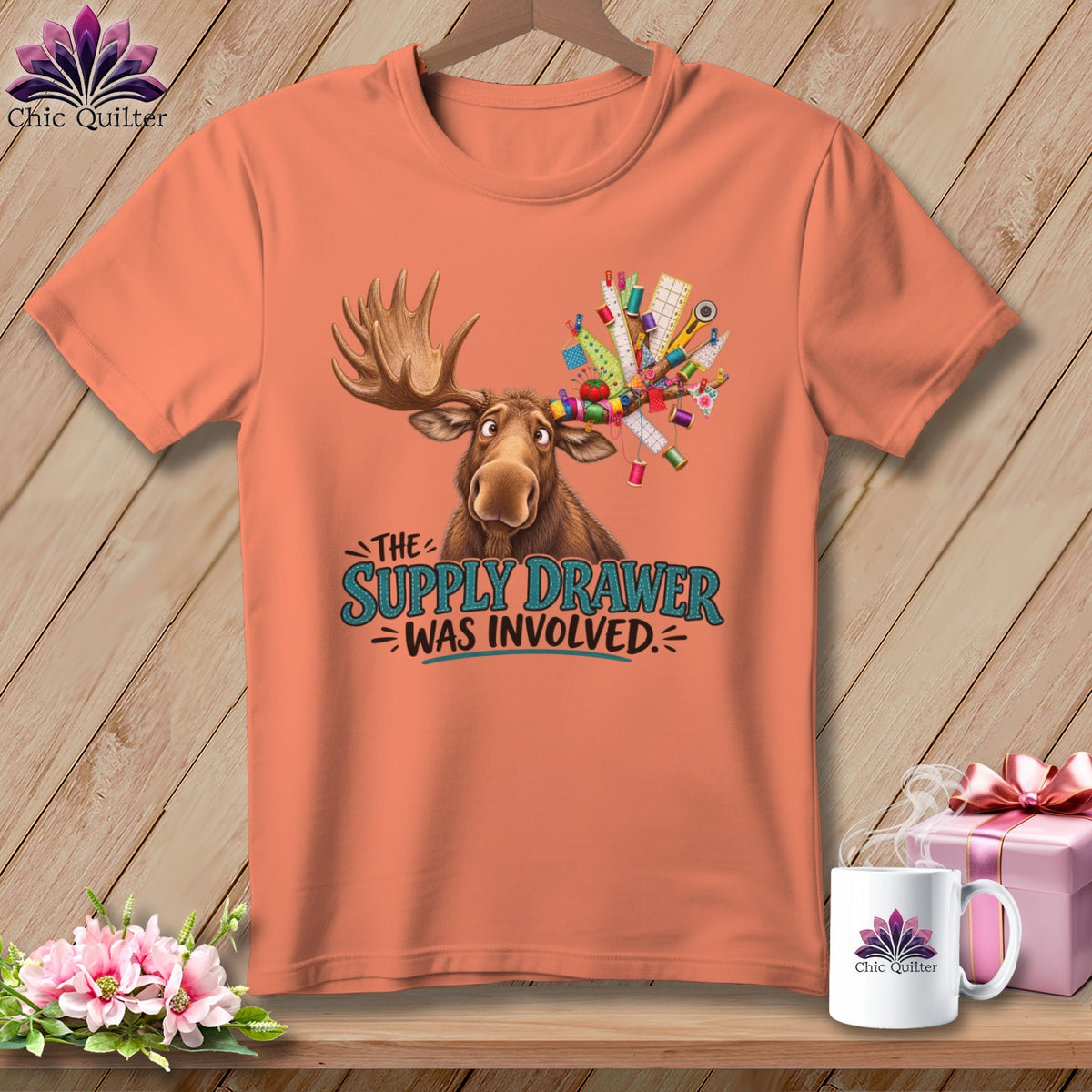 Peach t-shirt with a moose holding sewing supplies and text "The Supply Drawer Was Involved."