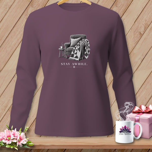 Purple long-sleeve shirt with a drawing of an armchair, quilt, and mug with "STAY AWHILE." text.