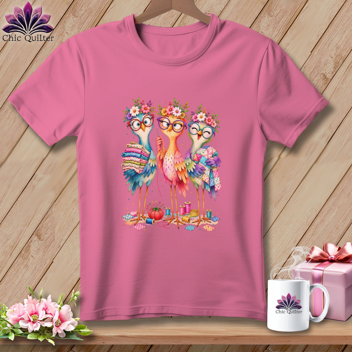 Pink t-shirt featuring an illustration of three colorful cartoon birds sewing, surrounded by crafting supplies.