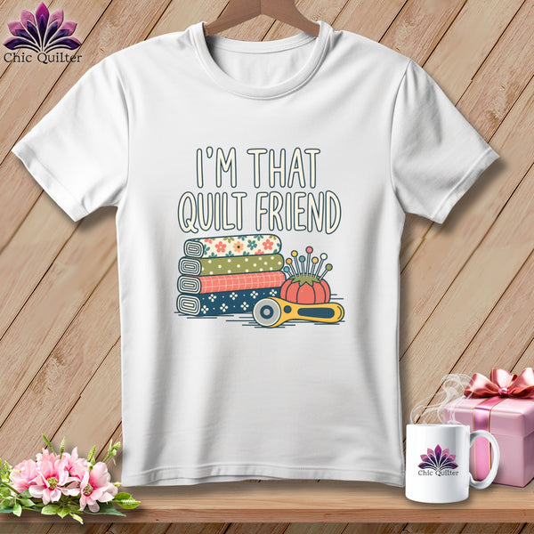 A white t-shirt features text reading I'M THAT QUILT FRIEND above fabric rolls, a pincushion, and a rotary cutter.