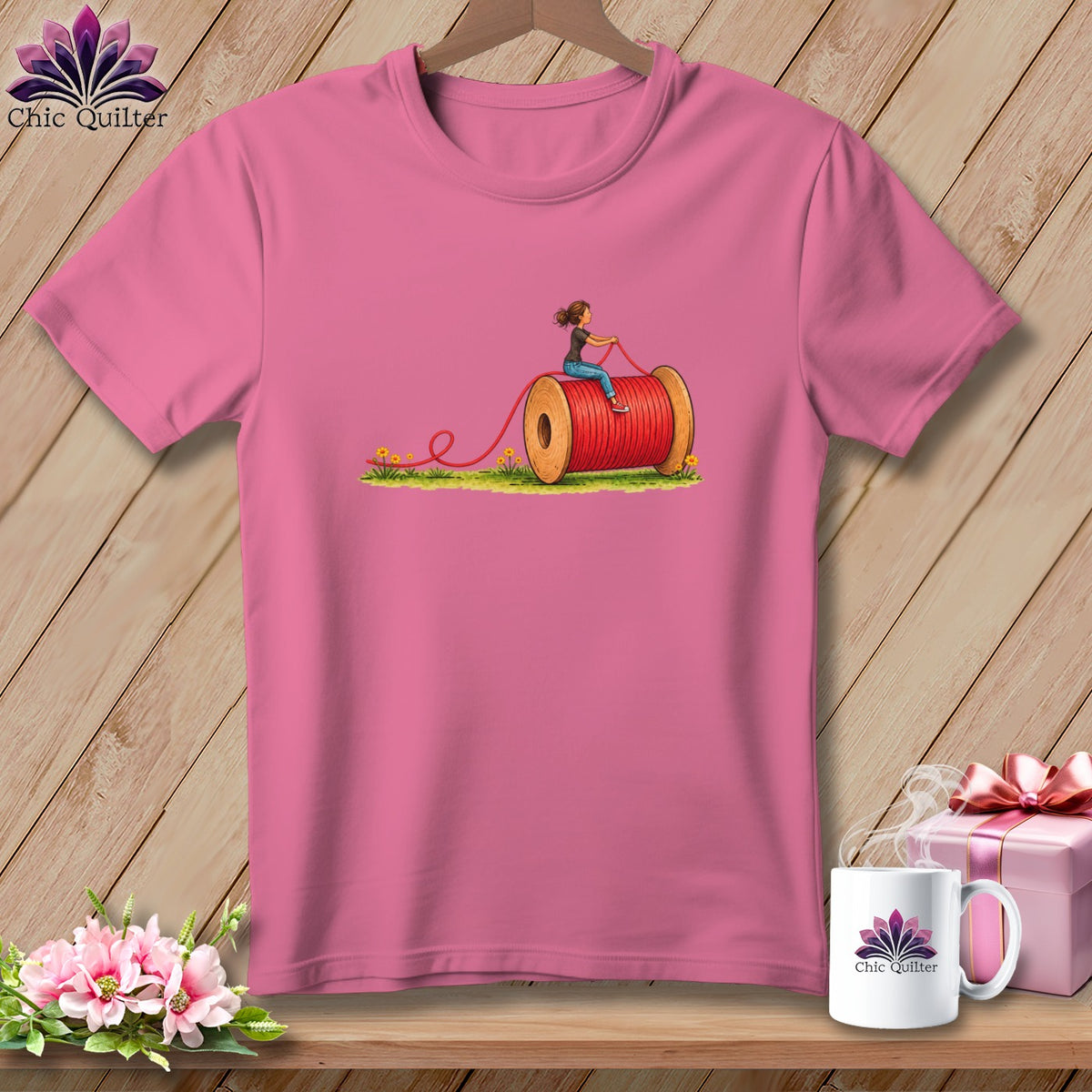 Pink short-sleeved t-shirt with a graphic of a woman riding a giant red thread spool on grass.