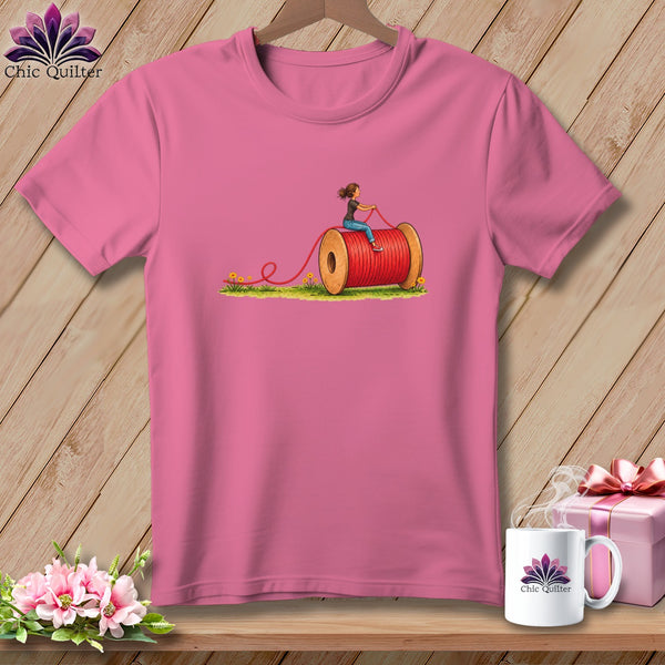 Pink short-sleeved t-shirt with a graphic of a woman riding a giant red thread spool on grass.