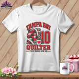 A white t-shirt features a graphic with a stack of quilts, a coffee mug, and text reading Tampa Bay Quilter First Down, Sew On!