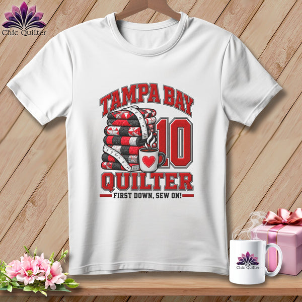 A white t-shirt features a graphic with a stack of quilts, a coffee mug, and text reading Tampa Bay Quilter First Down, Sew On!