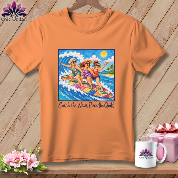 Orange t-shirt featuring a graphic of three women surfing on a patchwork quilt, with the text Catch the Wave, Piece the Quilt.