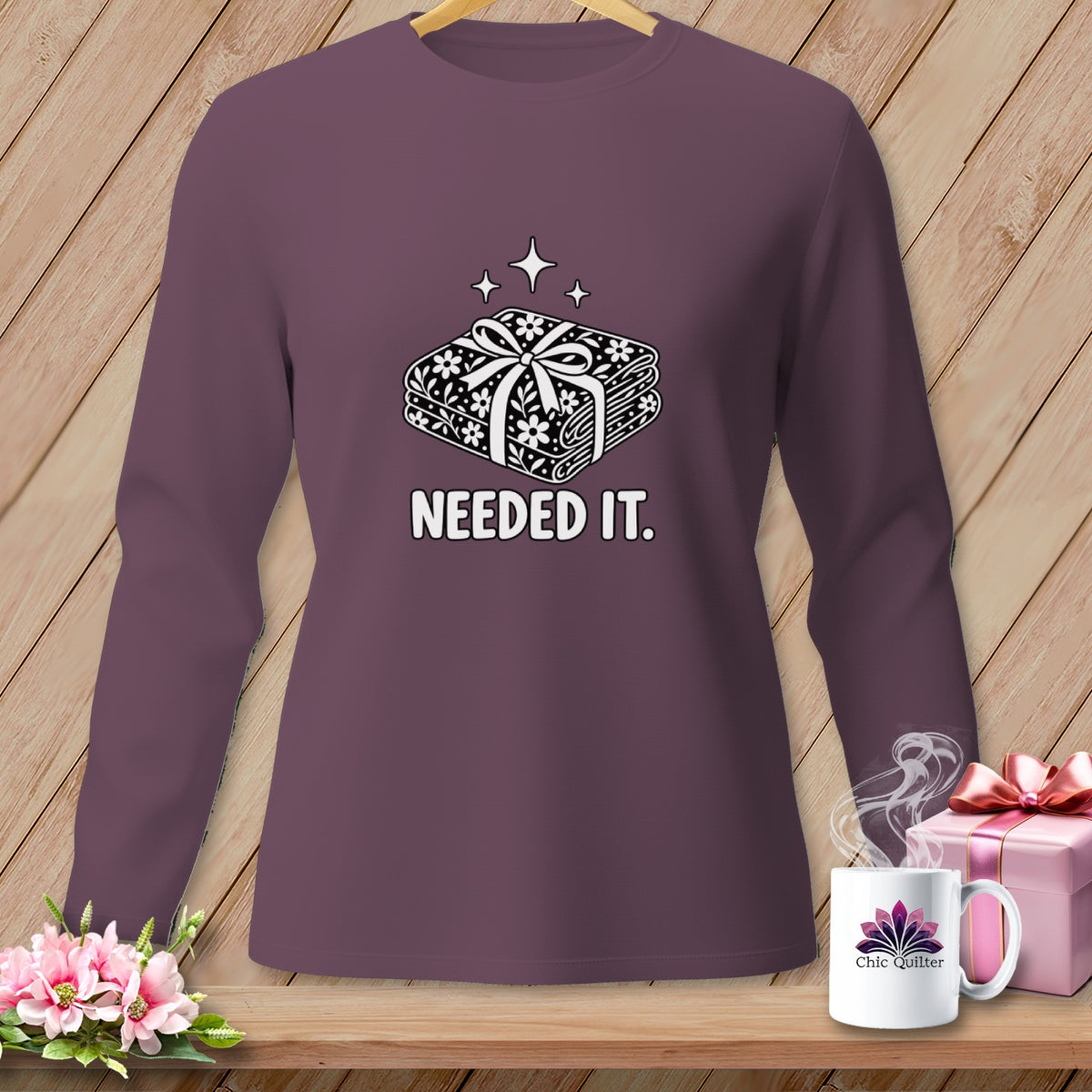 Purple long-sleeve shirt with a graphic of stacked fabric bundles and the text "NEEDED IT."