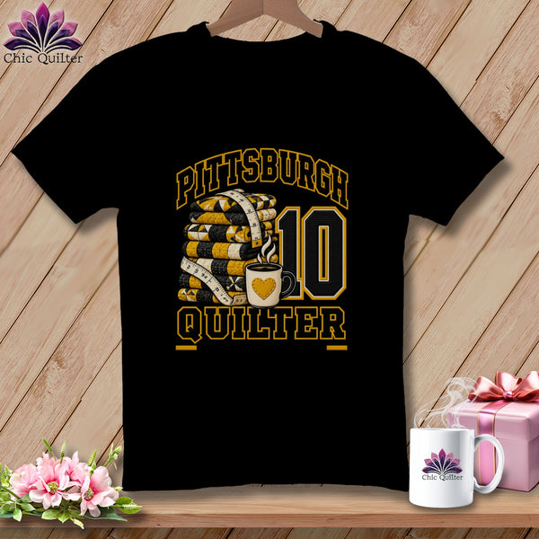 Black t-shirt featuring a Pittsburgh Quilter graphic with folded quilts, a measuring tape, coffee mug, and the number ten.