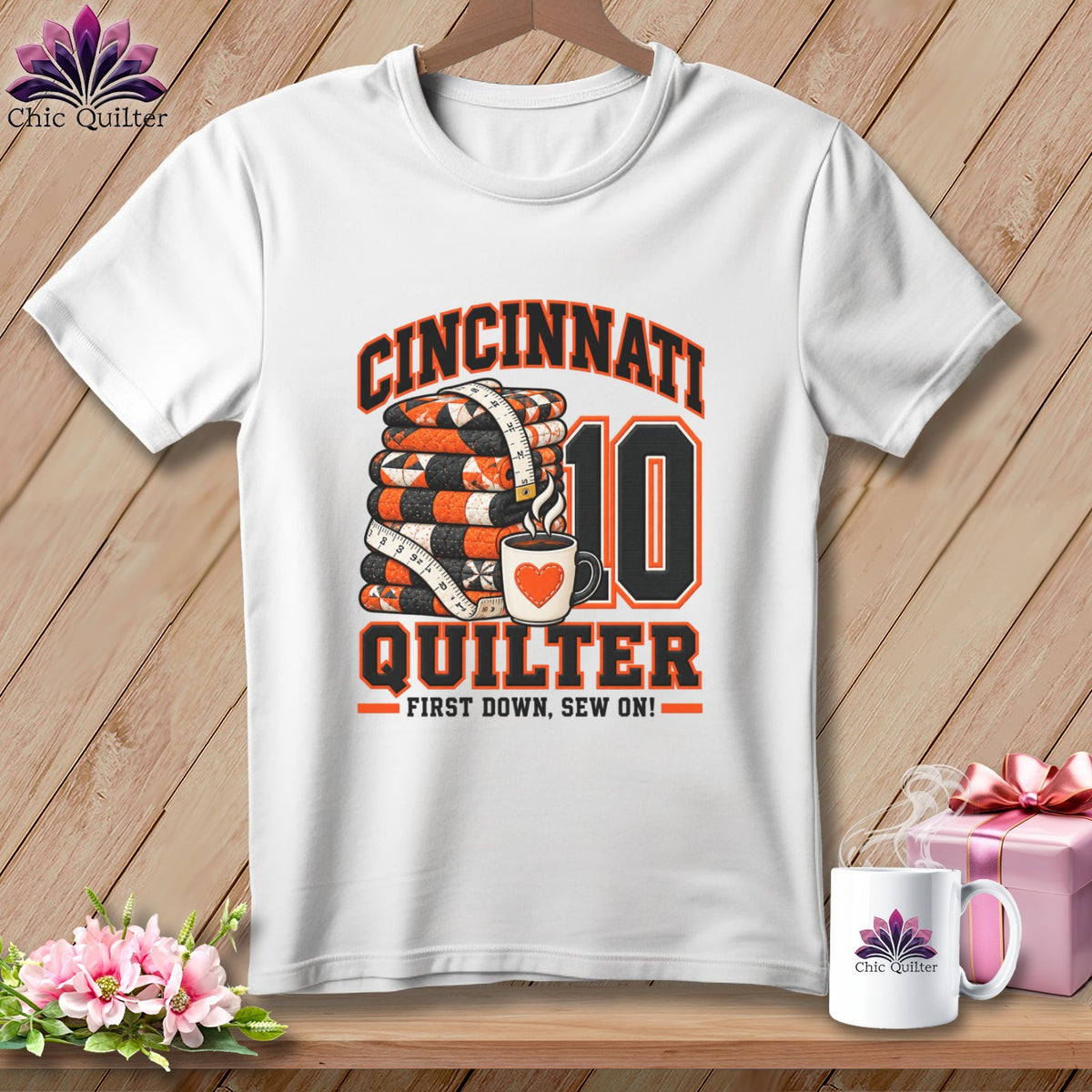 White t-shirt with a Cincinnati Quilter graphic featuring a stack of quilts, coffee mug, and text.