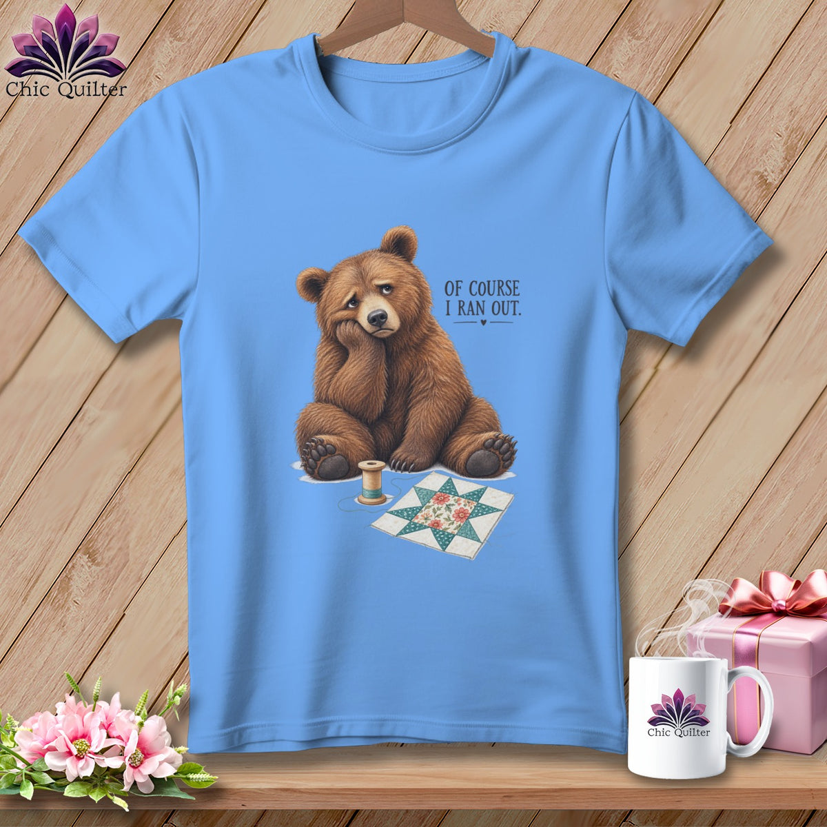Light blue t-shirt with a sad bear next to a quilt square and spool of thread.