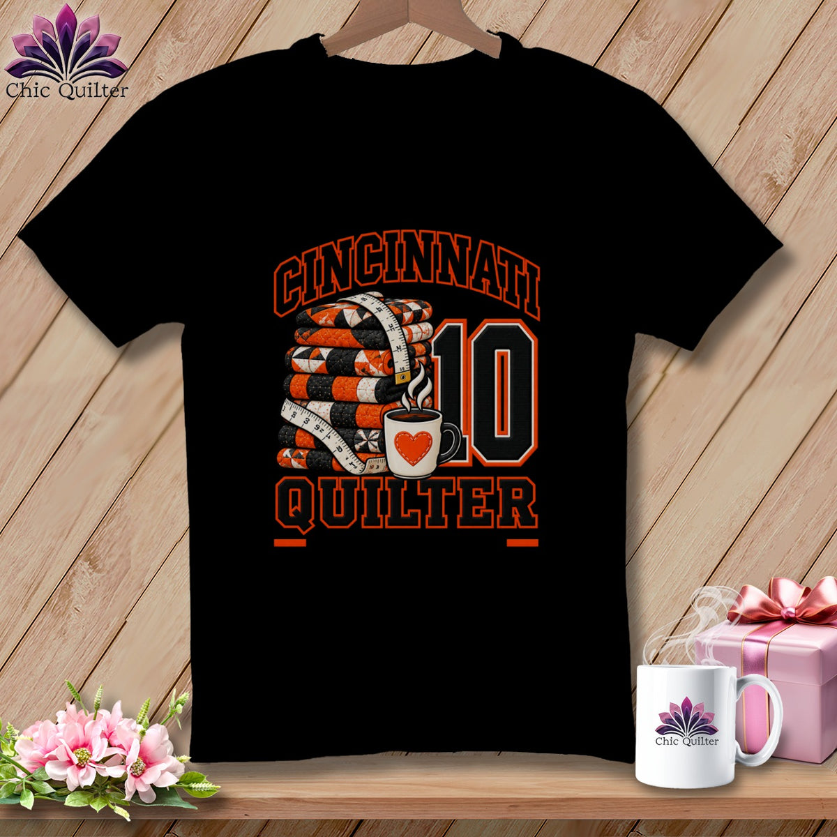 A black t-shirt featuring a graphic with a stack of quilts, a measuring tape, coffee cup, and Cincinnati Quilter text.