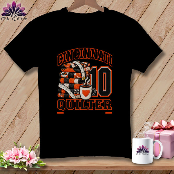 A black t-shirt featuring a graphic with a stack of quilts, a measuring tape, coffee cup, and Cincinnati Quilter text.