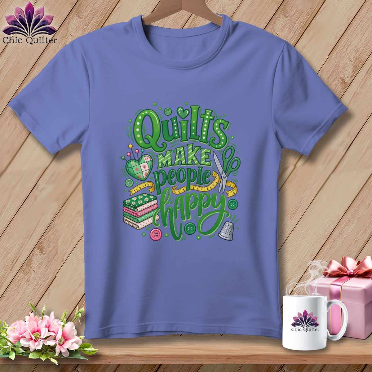 A periwinkle t-shirt on a wooden hanger features green text reading Quilts Make People Happy surrounded by sewing motifs.