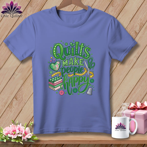A periwinkle t-shirt on a wooden hanger features green text reading Quilts Make People Happy surrounded by sewing motifs.