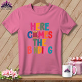 Pink t-shirt with multicolored text reading Here Comes the Binding and a spool of thread.