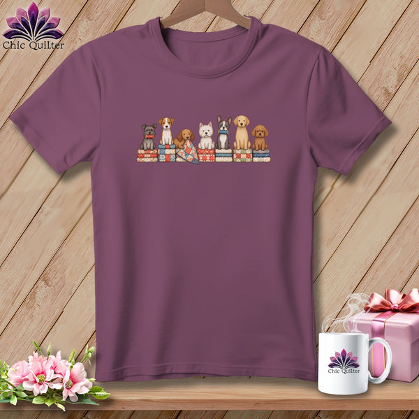 Mauve short-sleeve t-shirt hanging on a wooden wall, featuring a graphic of seven cartoon dogs sitting on stacked quilts.