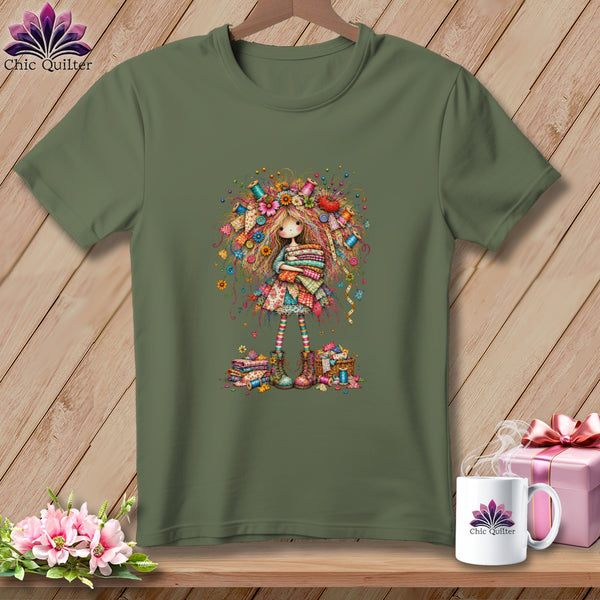 An olive green t-shirt featuring a whimsical illustration of a girl holding fabric stacks.