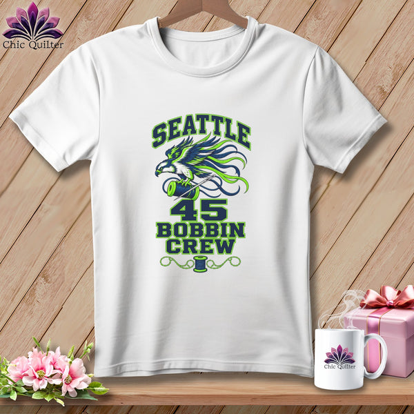 A white t-shirt hanging on a wooden background, featuring green and blue text that reads "Seattle 45 Bobbin Crew" alongside a bird and sewing imagery.