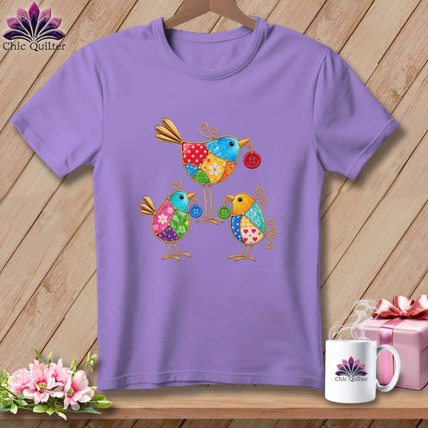 A lavender t-shirt featuring a colorful patchwork design of three stylized birds on a wooden background.