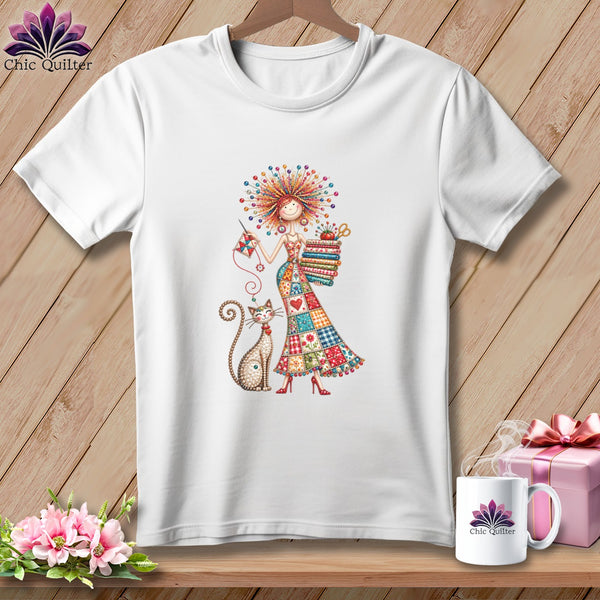 A white t-shirt hanging on a wooden background features a whimsical cartoon graphic of a woman holding a stack of colorful folded fabrics alongside a cat, with a matching mug and gift box on the shelf below.