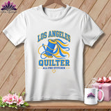 A white t-shirt hanging on a wooden background, featuring a Los Angeles Quilter graphic with a spool of thread and a needle.