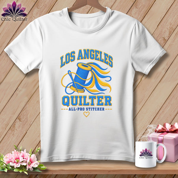 A white t-shirt hanging on a wooden background, featuring a Los Angeles Quilter graphic with a spool of thread and a needle.