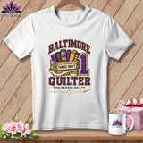 A white t-shirt featuring a Baltimore Quilter graphic with fabric rolls in a wooden crate.