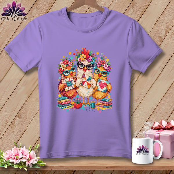 A purple t-shirt featuring a graphic of three cute chickens wearing glasses and floral crowns while holding quilts.