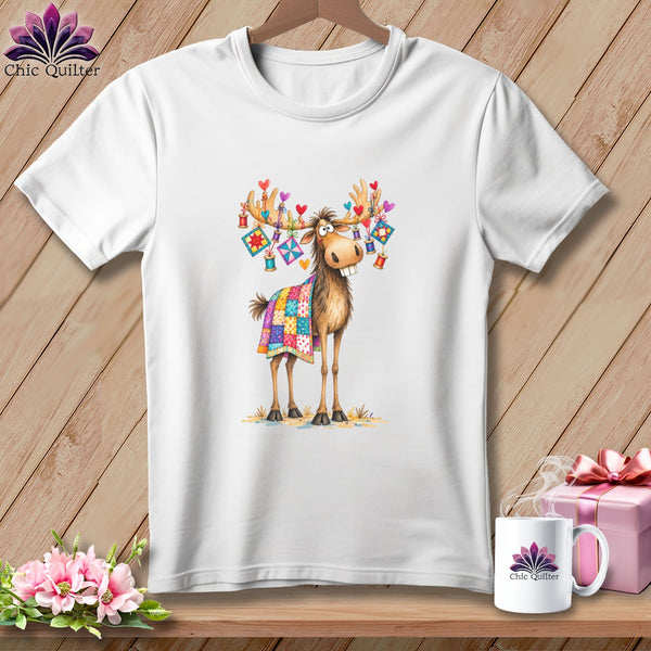 A white crewneck t-shirt featuring a whimsical moose graphic with a patchwork quilt and hanging heart decorations.